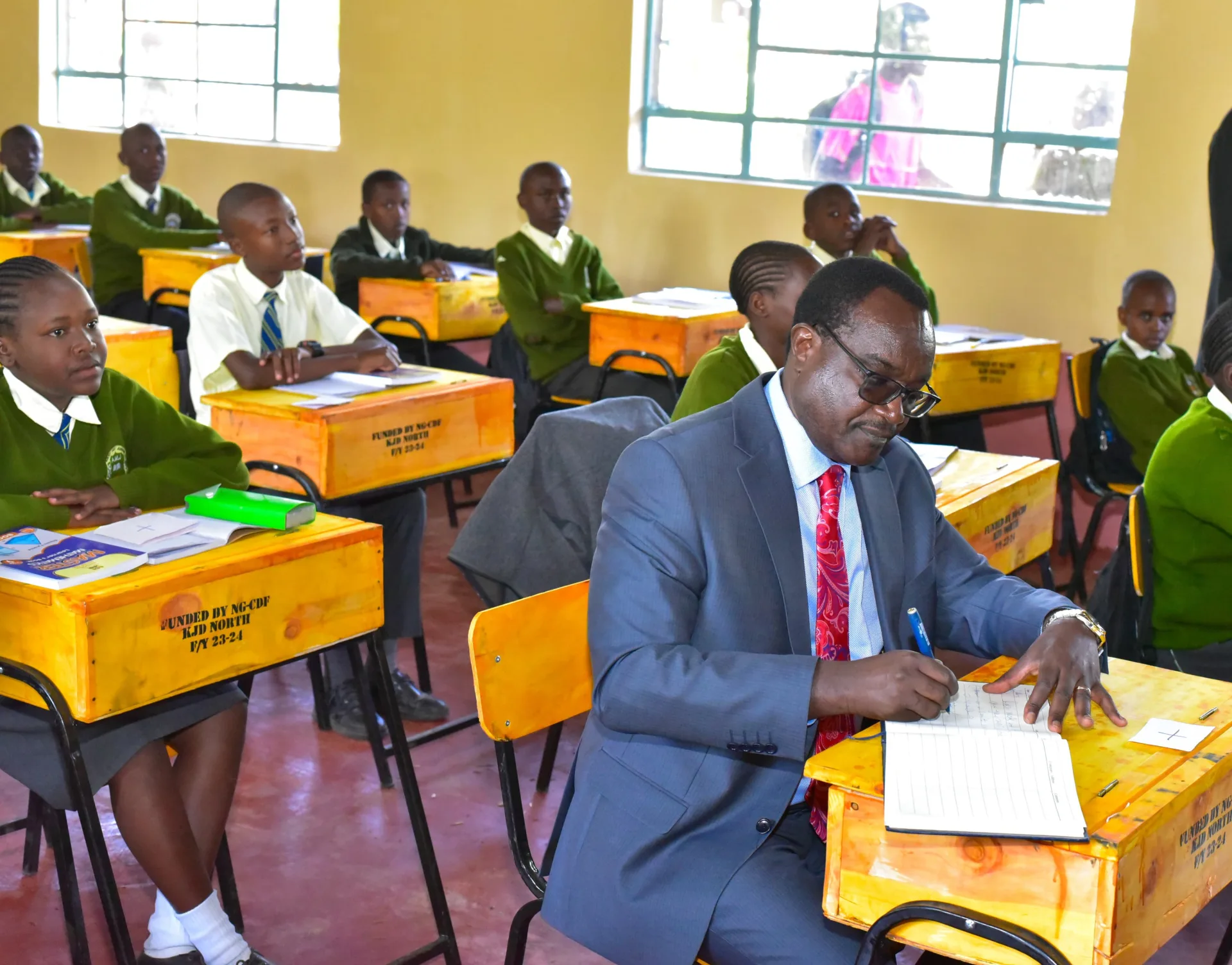 Education Cabinet Secretary Julius Ogamba writing during a school visit with learners seated on desks funded by NG-CDF in Kajiado North