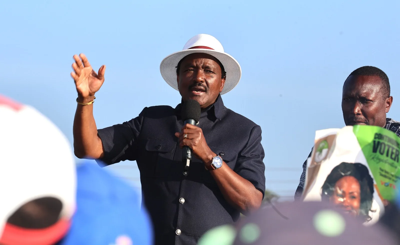 Kalonzo Musyoka speaking at a rally wearing white hat holding microphone