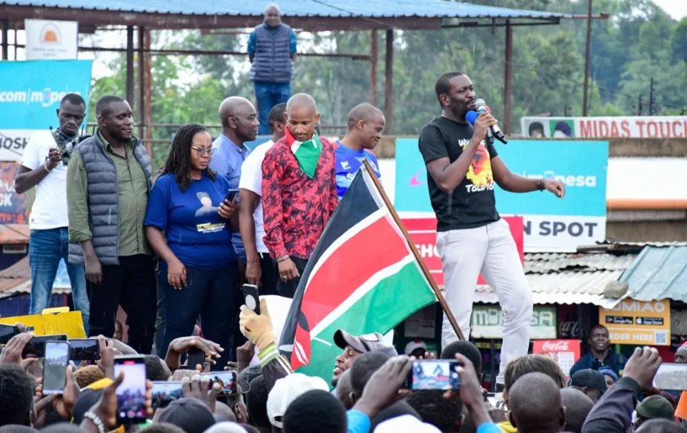 Nairobi Senator Edwin Sifuna speaking into microphone at Linda Mwananchi rally with Babu Owino Godfrey Osotsi and other leaders behind him crowd filming with phones Kenyan flag in foreground